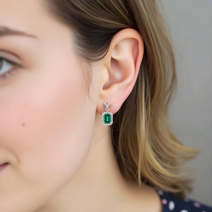 Model wearing Aeterna Jewelry emerald cut drop earrings, close-up ear view showing vivid green gemstones surrounded by sparkling white halo stones