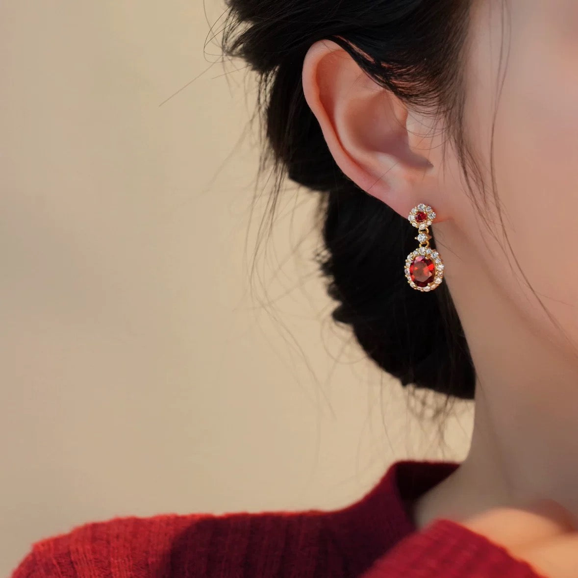 Model wearing Aeterna Jewelry vintage ruby halo drop earrings, elegant close-up ear view showing oval red gemstones in gold setting, soft indoor lighting